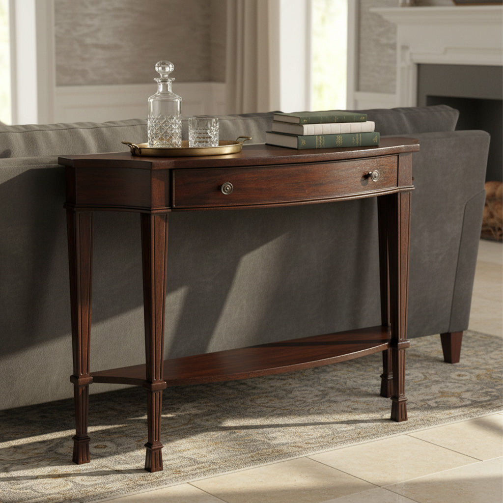 Breakfront curved mahogany sofa table with center drawer, antique brass knobs, and lower shelf behind gray sofa in living room