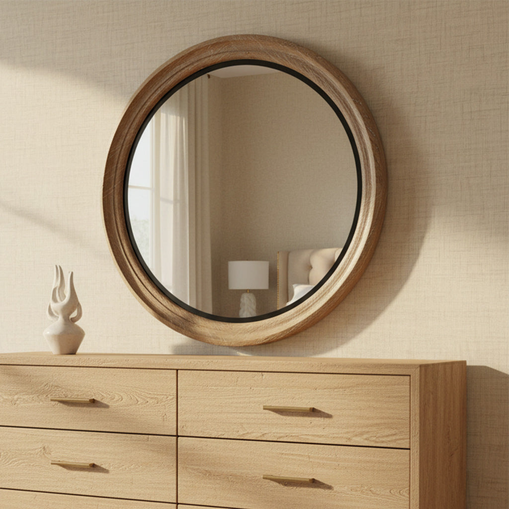 Round wall mirror with carved natural wood frame above light wood dresser next to white decorative sculpture in neutral room